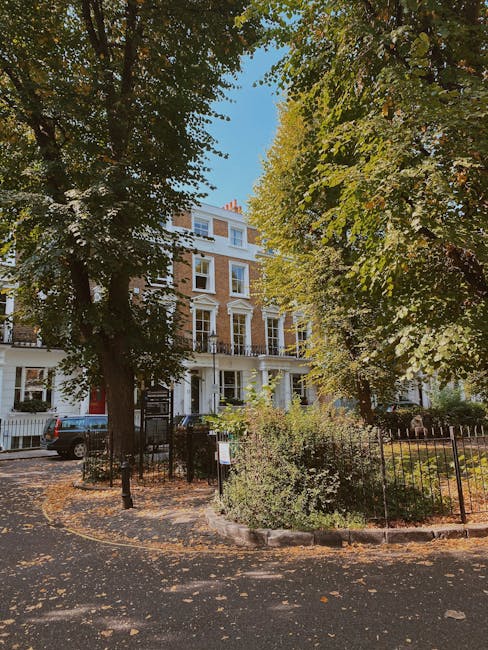 Photograph of a multi-story residential building on Broadhurst Gardens in West Hampstead, visible through large leafy trees with green and yellow foliage suggesting early autumn. The building is white with multiple large windows and small balconies on the upper floors, and a white entrance porch. A black metal fence surrounds a small front garden with shrubs and a pathway leading to the entrance. In front of the building, a paved pathway is partially covered with fallen leaves, and a parked dark-colored car is visible to the left. The scene is lit by bright daylight, highlighting the vibrant leaves and the building's facade. This setting encapsulates the typical environment for house removals and furniture transport services, with the street location suitable for loading and unloading items as part of a home relocation project. Man with Van West Hampstead may carry out furniture transport or packing and moving services in such residential areas.