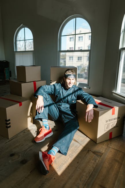 A young man with long hair and a headband, dressed in casual dark blue clothing and bright orange sneakers, sitting on a wooden floor inside a room with large arched windows. He is surrounded by multiple packed cardboard boxes sealed with red and black tape, arranged in a semi-circle around him. The sunlight streaming through the windows illuminates the scene, highlighting the packing materials and the interior space. The setting suggests a home relocation or packing process as part of a moving service, with the presence of boxes indicating preparation for transportation. This image demonstrates the packing phase of a furniture and household item move, relevant for companies like Man with Van West Hampstead specializing in house removals and logistics for local moves in NW6, West Hampstead.