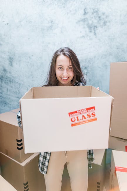 A young woman with long brown hair, smiling and looking directly at the camera, is standing indoors against a light blue textured wall. She is holding an empty cardboard moving box with a red label that reads 'FRAGILE GLASS' on the front. Surrounding her are several other packed cardboard boxes of various sizes, some with black tape reinforcement and others with red or white labels, indicating preparations for a house or student move. The boxes are placed on a smooth, light-colored floor, with a partially visible closed cardboard box to her right. This scene captures a moment of packing or unpacking, typical of home relocation or furniture transport processes, executed by a professional moving service like Man with Van West Hampstead. The lighting is bright and even, emphasizing the clean, organized environment suitable for logistical and moving activities.