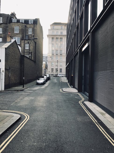 A narrow residential street in NW6, West Hampstead, with parked cars along the curb on the left side and a row of multi-storey brick buildings on either side. The street surface is asphalt with double yellow lines marking the edge of the roadway. On the right side, a modern building with dark-colored metal cladding and a rolling metal shutter door is visible, indicating a loading area. The perspective shows the street narrowing towards a larger, white stone building in the background. The scene is well-lit with natural daylight, reflecting typical urban conditions suitable for house removals and furniture transport processes, with space for loading and unloading during home relocation or packing and moving activities, supported by local removal specialists such as Man with Van West Hampstead.