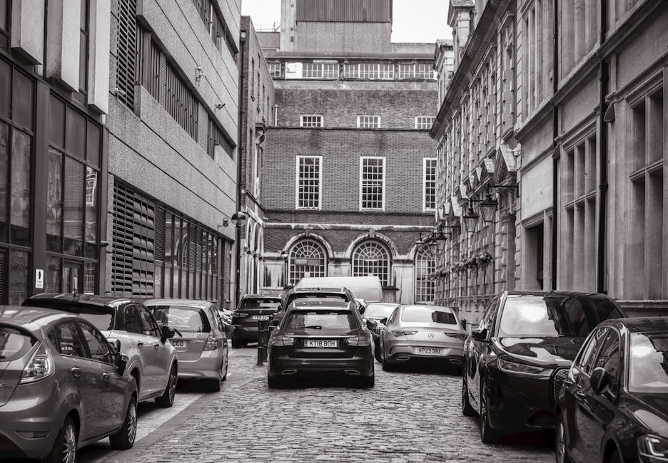 A narrow cobblestone street in West Hampstead lined with parked cars on both sides, including hatchbacks, sedans, and an estate vehicle, with some vehicles partially blocking the sidewalk. On the left, a modern building features large windows with dark frames, while the right side showcases a historic-looking building with classical architectural details and window boxes. In the background, there is a brick building with multiple tall, narrow windows and a decorative archway. The scene is in black and white, emphasizing the tight urban environment typical for home relocation or furniture transport activities, as part of a professional move involving careful vehicle and furniture handling in confined city streets. Man with Van West Hampstead's removals service may be involved in navigating parking and street access challenges for furniture transportation and loading process.