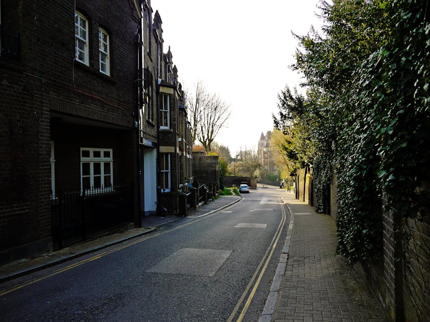 A narrow residential street in NW6, West Hampstead, with parked cars along the curb on the left side and a row of multi-storey brick buildings on either side. The street surface is asphalt with double yellow lines marking the edge of the roadway. On the right side, a modern building with dark-colored metal cladding and a rolling metal shutter door is visible, indicating a loading area. The perspective shows the street narrowing towards a larger, white stone building in the background. The scene is well-lit with natural daylight, reflecting typical urban conditions suitable for house removals and furniture transport processes, with space for loading and unloading during home relocation or packing and moving activities, supported by local removal specialists such as Man with Van West Hampstead.