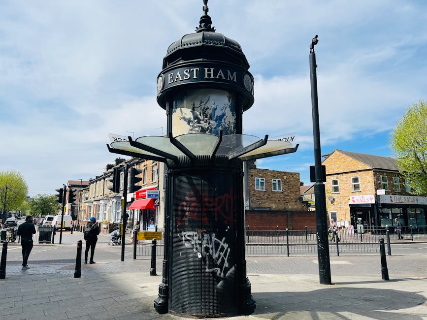 A black, cylindrical clock tower with a domed top displaying the East Ham name is situated at the corner of a busy urban street. The tower features a round face with decorative elements, and the station name is prominently shown on a curved signage panel near the top. The surrounding environment includes a paved sidewalk, several pedestrians walking nearby, and light traffic with parked and moving vehicles. To the right, there is a small commercial building with signage for a nail salon, and a tree with green leaves is visible in the background under a partly cloudy sky, suggesting daytime. The scene captures a typical street view, suitable for discussing transportation or urban infrastructure related to home relocation or furniture transport services offered by Man with Van West Hampstead.