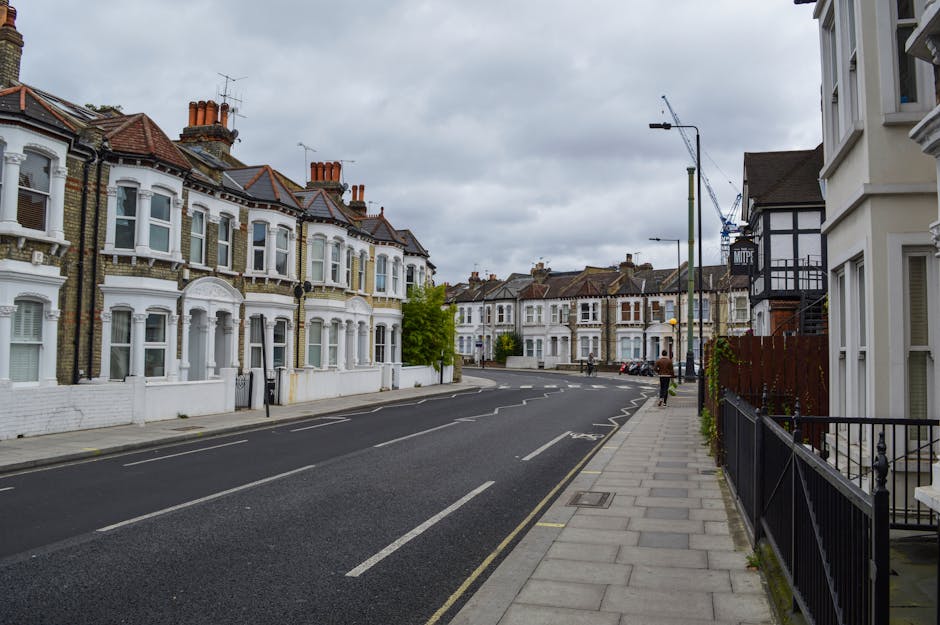 An empty residential street in West Hampstead with row houses on the left featuring white facades, bay windows, and red-tiled roofs. The pavement runs along the right side, bordered by a black iron fence and front gardens. A streetlamp and parked cars are visible further down the road. Overhead, the sky is cloudy, indicating overcast weather. The scene, taken during daylight, depicts a typical suburban neighbourhood ready for home relocation or moving services, with no people or active loading visible. This image exemplifies the environment in which a professional removals company, such as Man with Van West Hampstead, might operate during furniture transport or packing and moving tasks in NW6.
