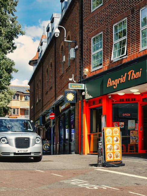 The image depicts a street scene in West Hampstead with a brick building housing several shops, including a Thai restaurant with a red facade and an open sign in the window. A silver vehicle is parked on the left side of the street. In front of the shops, there are cardboard boxes and furniture wrapped in plastic and fabric coverings, indicating the process of home relocation or furniture transport. A hand truck or trolley is visible, being used to carry or transport packing materials or furniture onto the pavement outside the retail units. The scene is well-lit with natural daylight, and the background shows additional buildings, trees, and a partly cloudy sky. This setting is relevant to the context of house removals, packing, and moving services offered by Man with Van West Hampstead, exemplifying typical activities involved in loading and unloading during a residential move in the NW6 area.