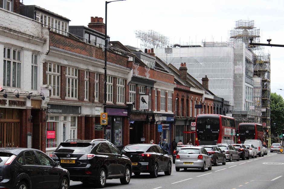 An empty residential street in West Hampstead with row houses on the left featuring white facades, bay windows, and red-tiled roofs. The pavement runs along the right side, bordered by a black iron fence and front gardens. A streetlamp and parked cars are visible further down the road. Overhead, the sky is cloudy, indicating overcast weather. The scene, taken during daylight, depicts a typical suburban neighbourhood ready for home relocation or moving services, with no people or active loading visible. This image exemplifies the environment in which a professional removals company, such as Man with Van West Hampstead, might operate during furniture transport or packing and moving tasks in NW6.